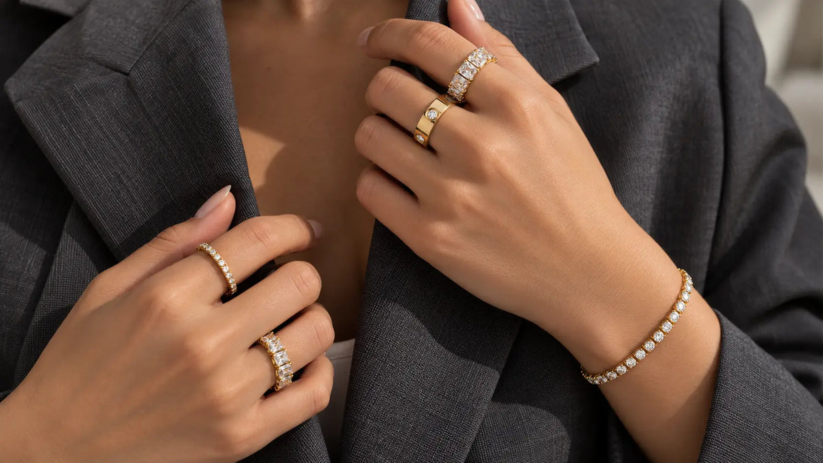 Kombinierte goldene Ringe mit Steinen und Tennis-Armband an den Händen vor einem grauen Blazer