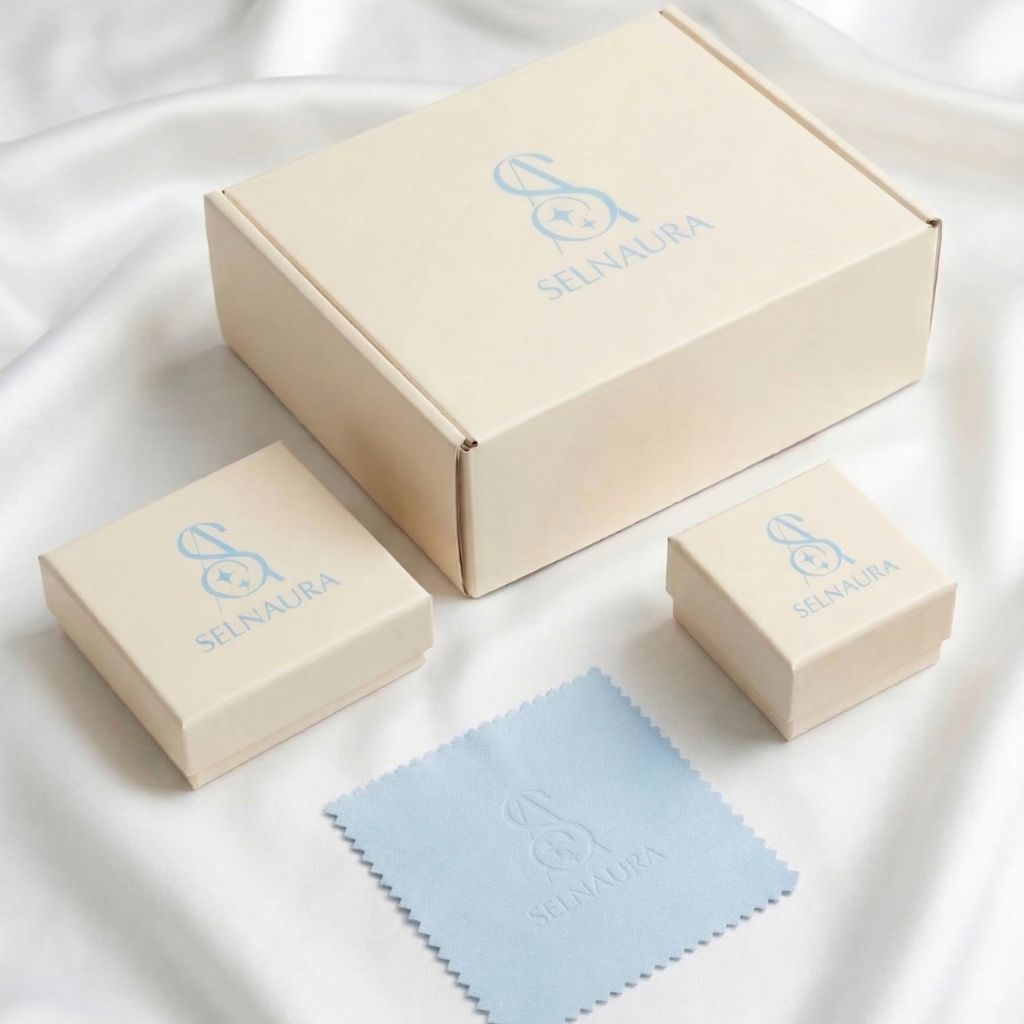 Selnaura Verpackungsset bestehend aus cremefarbener Versandbox, zwei Schmuckboxen in verschiedenen Größen und einem hellblauen Poliertuch mit Selnaura-Logo auf weißem Satin