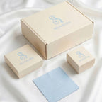 Selnaura Verpackungsset bestehend aus cremefarbener Versandbox, zwei Schmuckboxen in verschiedenen Größen und einem hellblauen Poliertuch mit Selnaura-Logo auf weißem Satin